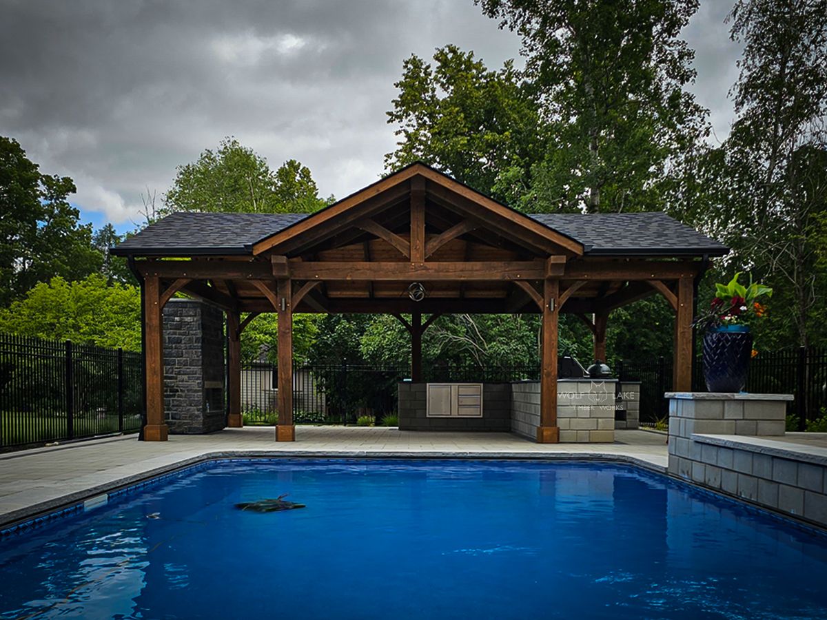 A wooden pavilion with a dark roof next to a swimming pool, featuring an outdoor kitchen and seating area.